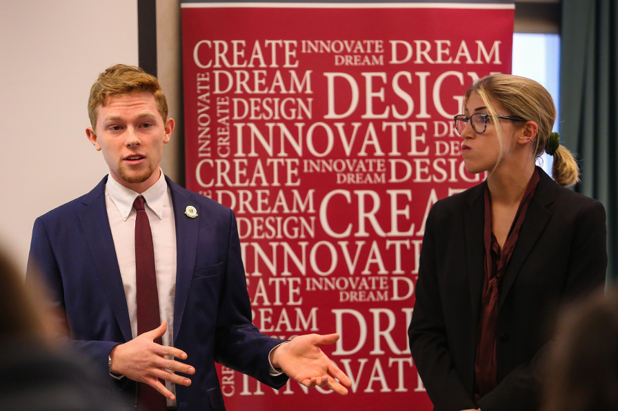 Students in business attire make a presentation in front of a backdrop banner stamped with words that include: Create. Dream. Innovate. Design.