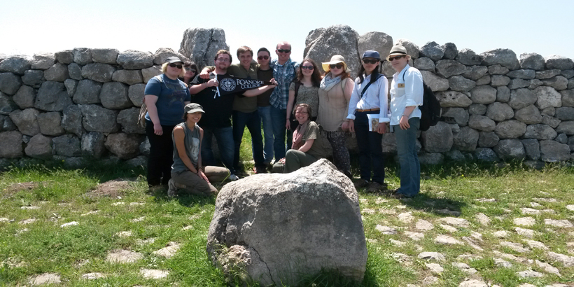 Students and faculty pose for a group photo next to a tall wall made from large stones