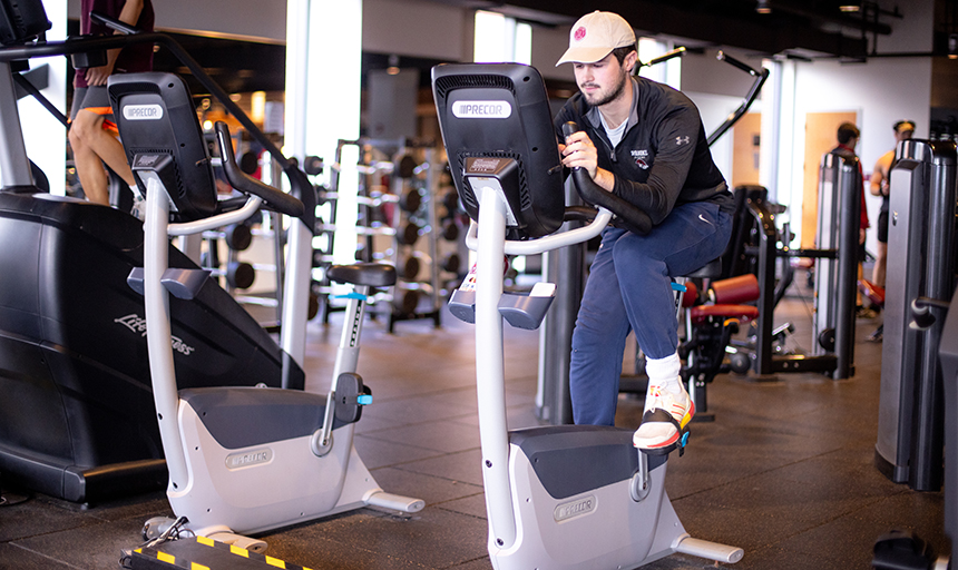 Young man in black pullover and tan hat rides an exercise bike in a fitness center with other exercise equipment in the background.