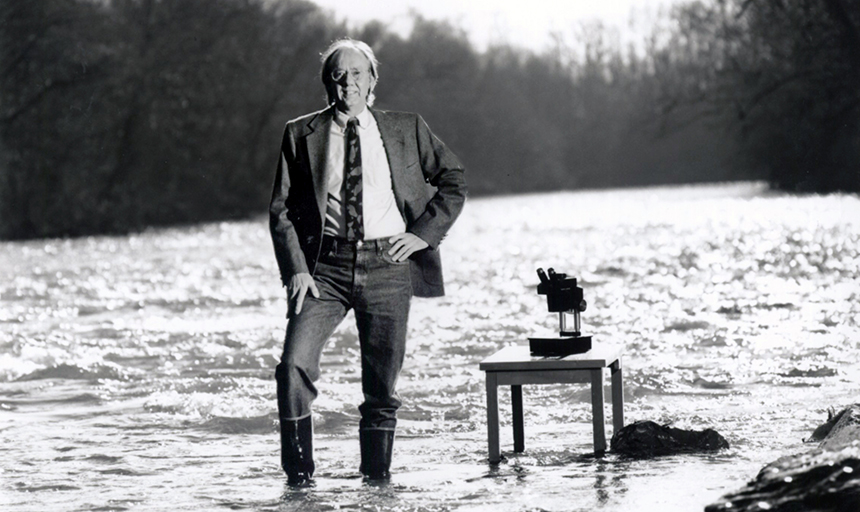 Black-and-white photo with Professor Bob Jenkins standing in the center of a river in suit and tie next to a small table with a microscope on top.