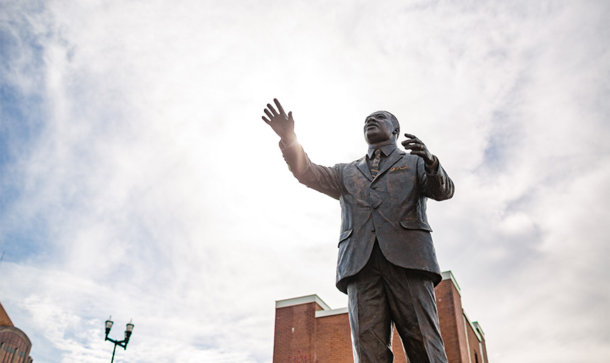 The sun shines behind a bronze statue depicting Martin Luther King Jr. with arms outstretched
