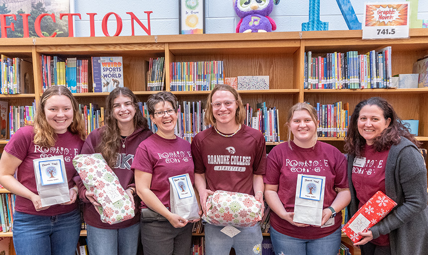 Professor McCutcheon and student volunteers with the Toy Like Me at Roanoke College club smile and hold presents while posing for a photo during a toy delivery in the Virginia Heights Elementary School library