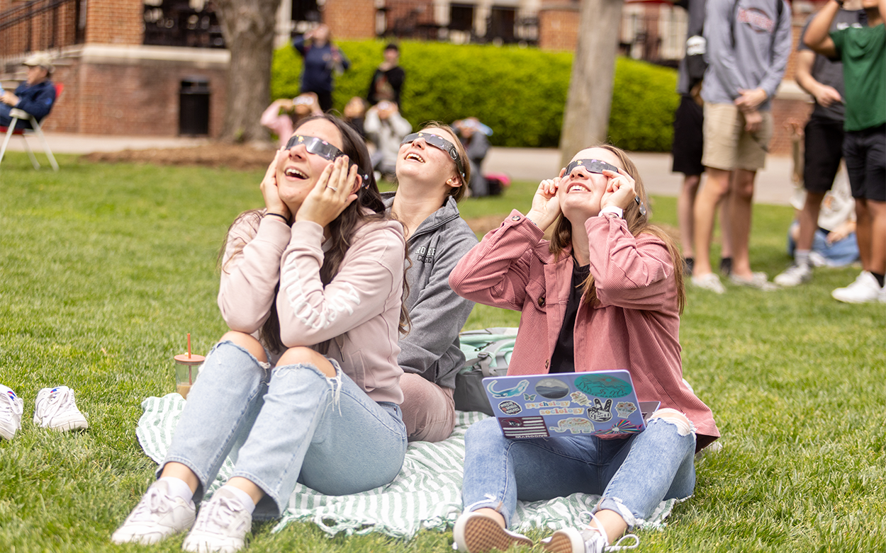 Three students wearing eclipse glasses look toward the sky