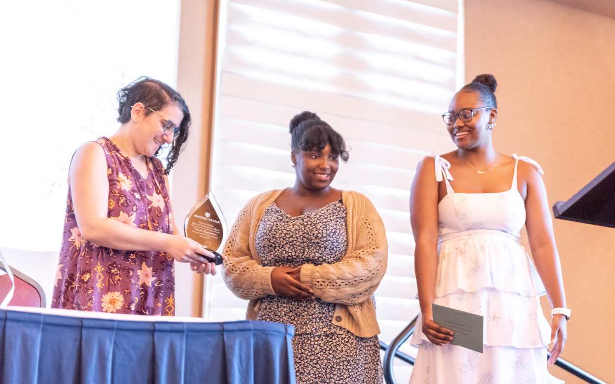 A woman looks down at an award while smiling after receiving it from two student presenters on stage