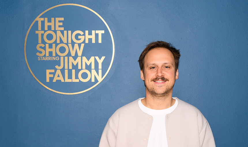 Brooks Allison stands, smiling, in front of a blue backdrop bearing the logo for "The Tonight Show with Jimmy Fallon"