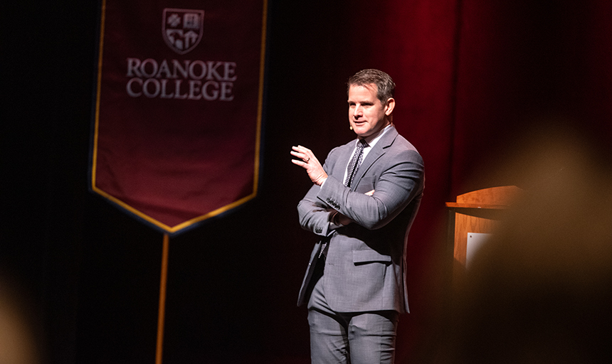 Adam Kinzinger, wearing a gray suit, gestures with one hand as he talks to the audience from the Olin Theater stage, and there is a maroon Roanoke College banner in the background.