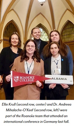 Four students, one holding a Roanoke College pennant and one holding an Albania sign, smile for a group photo with their faculty advisor at a conference