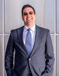 Man in dark suit, blue tie and tinted glasses smiles at camera in front of silver-paneled building.