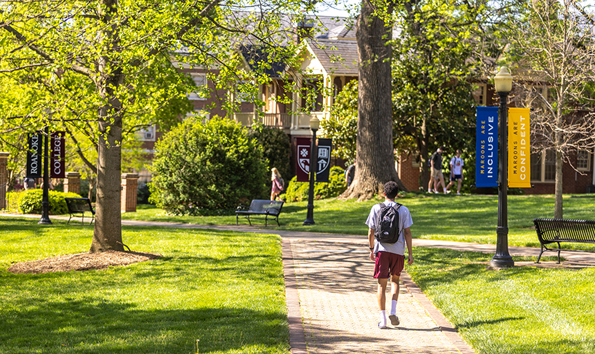 Student with a red backpack walks down a brick sidewalk with red brick campus buildings in the background.