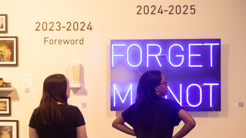 Students in an art gallery gaze at a neon sign that reads: Forget Me Not