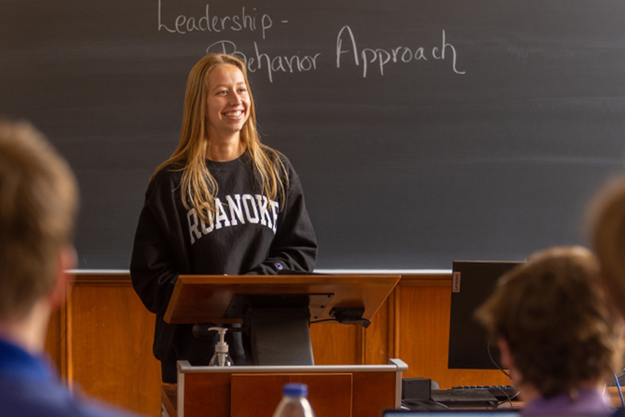 A student wearing a Roanoke sweatshirt stands in front of a chalkboard with the words Leadership Behavior Approach written on it