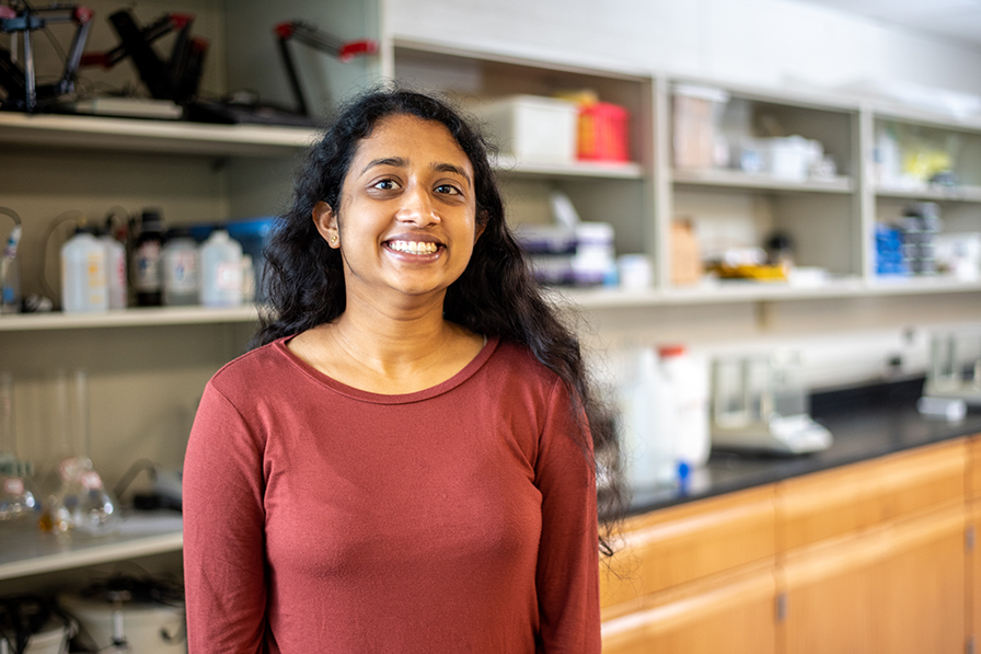 Kavya stands in a red shirt in a lab with shelves holding lab equipment behind her.