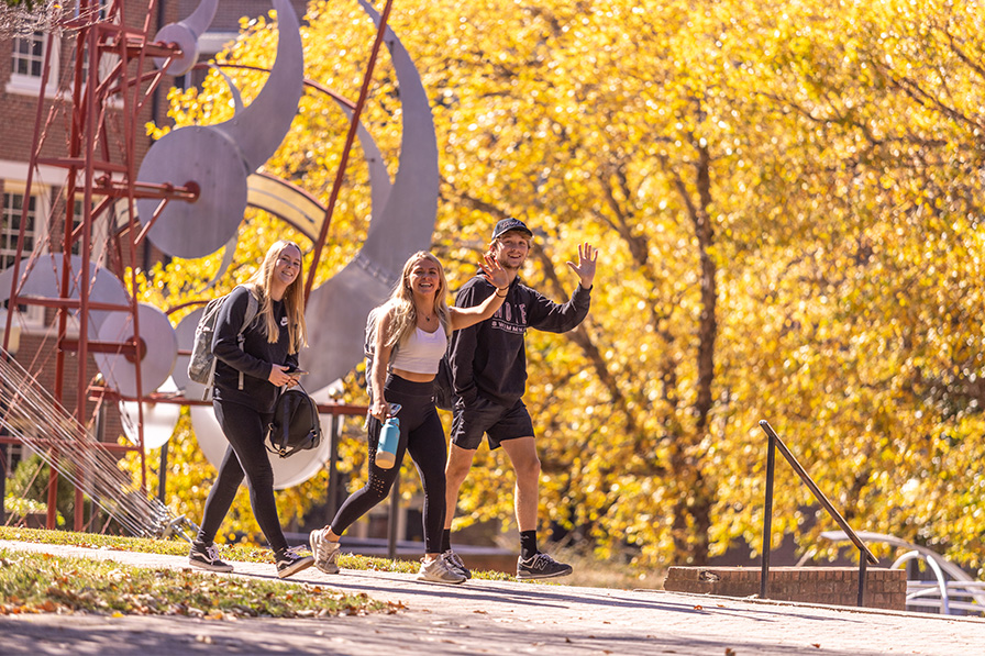 Students smile and wave at someone while walking across the quad against a background of autumn foilage