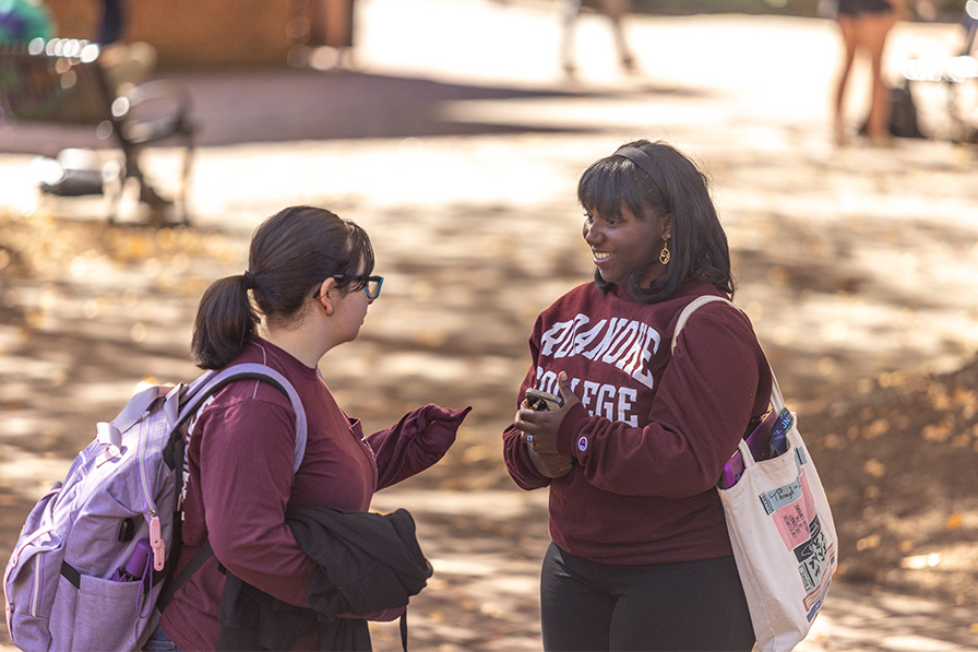 A student wearing a Roanoke sweatshirt smiles and greets another student on the quad