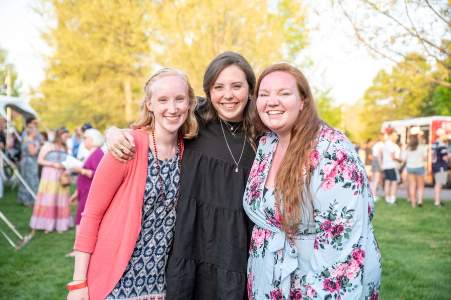 Three female alumni stand on the Back Quad with their arms around each other, looking at the camera and smiling.