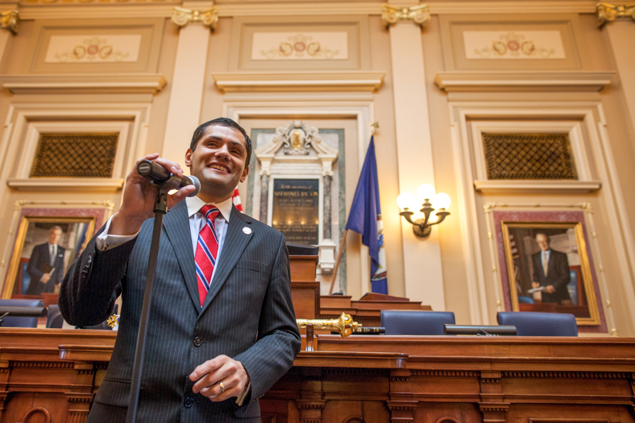A man in a dark suit and red tie stands in what appears to be a legislative office with a huge wooden desk and flag of Virginia behind him.