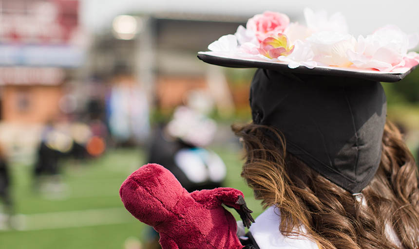 A graduate with flowers decorating her cap and a Rooney stuffed animal on her shoulder facing forward at commencement