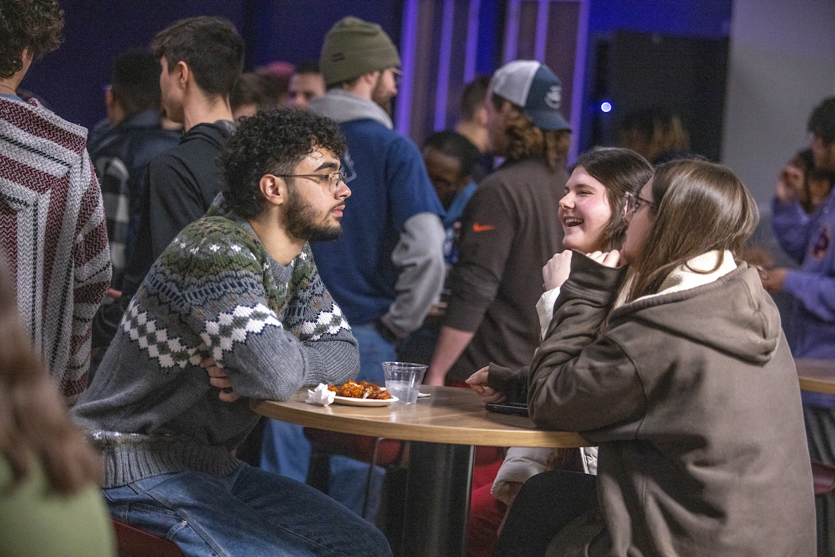 Three students sitting at a table eating wings with other students mingling behind them