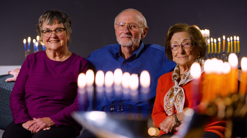 Helga Morrow, Arye Ephrath and Regine Archer sit together for a photo on a filming set dotted by lit menorahs.