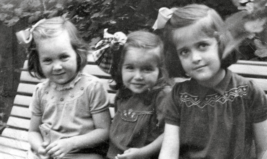 A black-and-white photo of three young girls sitting together on a park bench in 1940s Europe