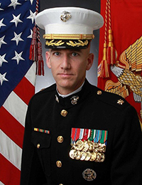 Man in a marine Corps uniform with many ribbons and medals on his chest poses with an American flag and a Marine Corps flag in the background.