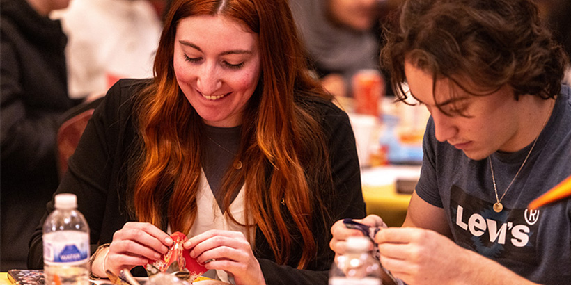 A student seated at a table smiles while folding paper to make an origami figure