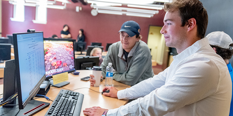 Three students, one leaning forward while holding a coffee cup, gather around a computer as their experiment results come in