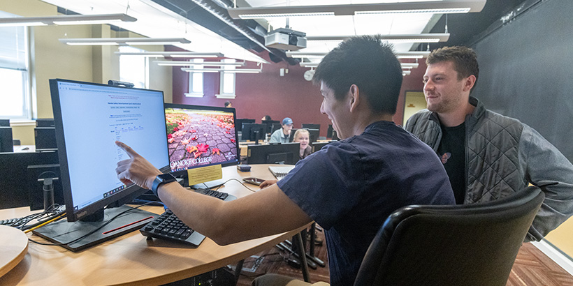 Two students, one reaching out his hand to point at a computer screen, discuss their team's experiment results