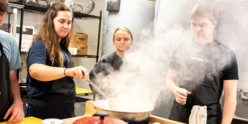 Three students, one holding a large spoon, watch over a pan that is emitting a large plume of steam