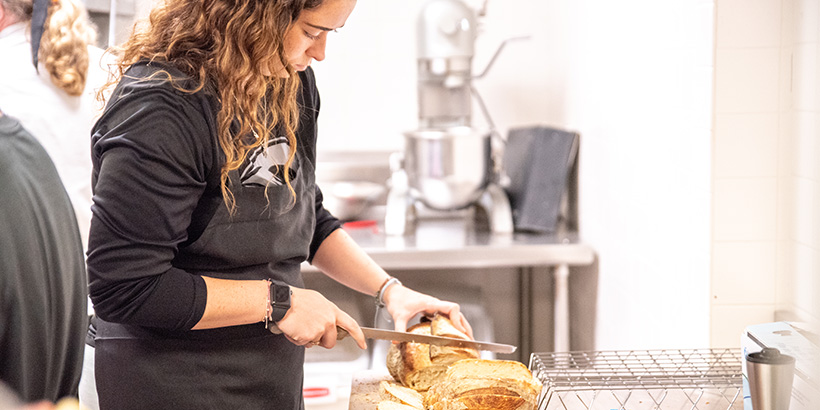 Madison Filson wields a long bread knife as she slices a loaf on a cutting board in a campus kitchen