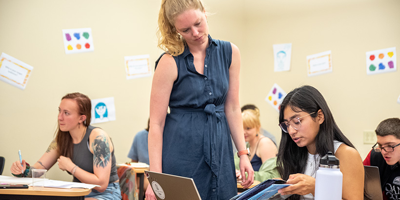 A teacher (standing) talks with a student (seated) while reviewing a book