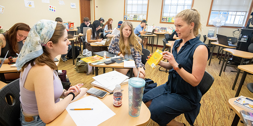 A teacher holds up a book while explaining a point to two students seated at desks in a classroom