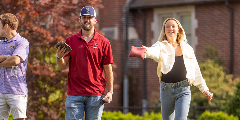 Close-up shot of two students, one about to release a throw, who are competing together on a cornhole team