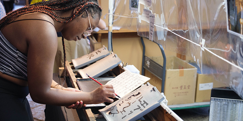 A student leans forward while wielding a calligraphy brush to create delicate lettering