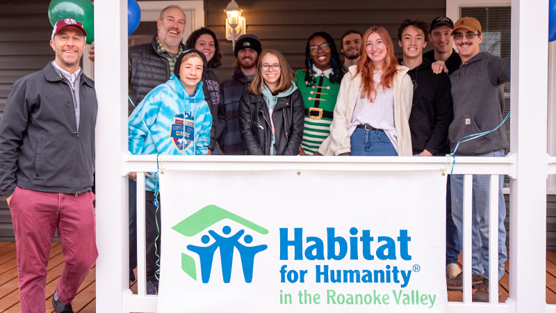 Keshia Jones and Roanoke College students, staff smile for a group photo on a porch draped with a Habitat for Humanity banner