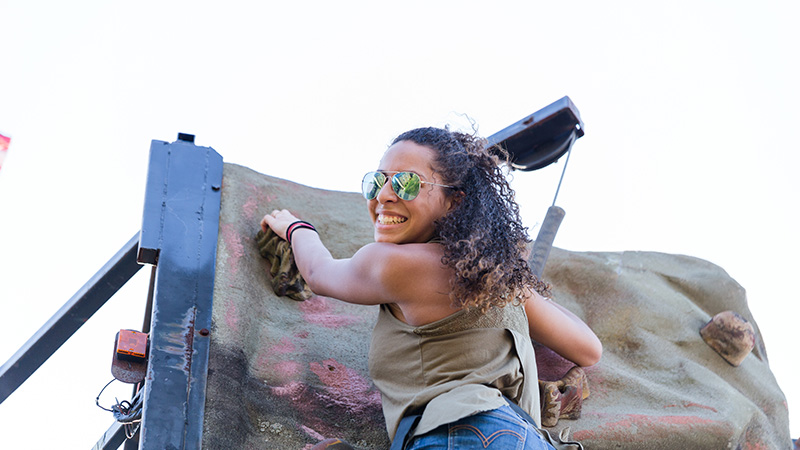 A woman wearing sunglasses smiles after reaching the top of an outdoor climbing wall