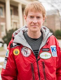 REd-headed man in a red jacket with adventure patches on it poses for a photo outdoors with a brick building in the background.