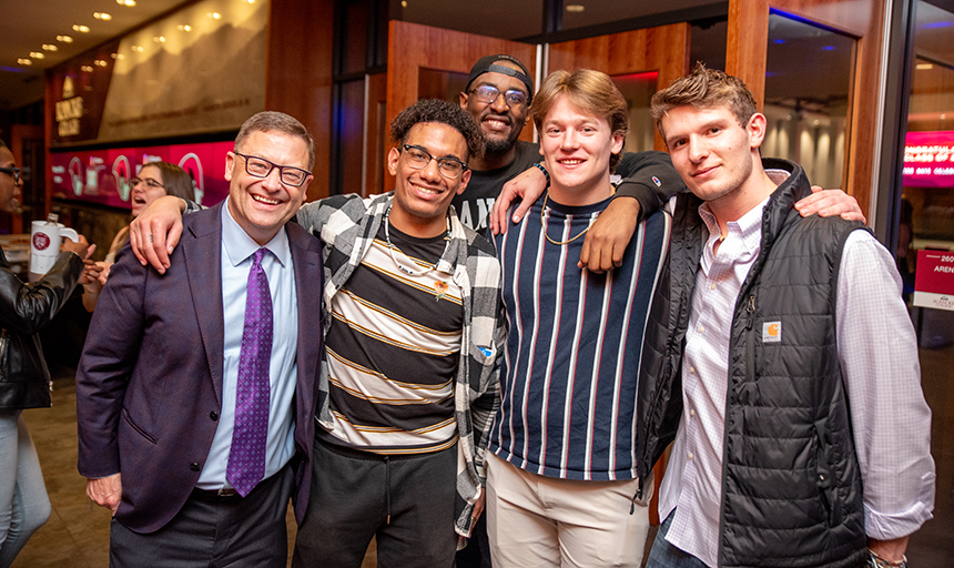 Man in a suit and tie poses for picture with four male college students, and all are smiling