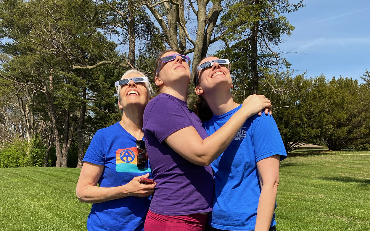 Three people stand together and look toward sky while wearing eclipse glasses