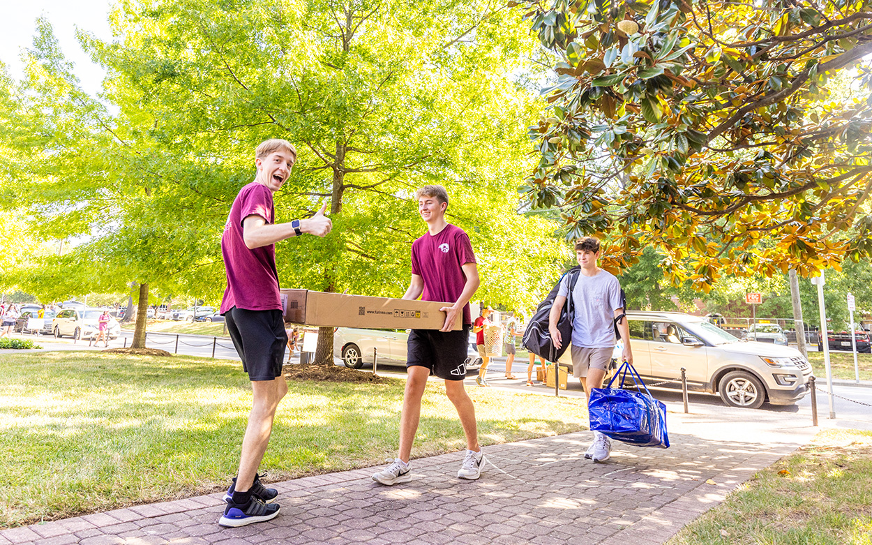 A student gives a thumbs up while helping carry a large box into a residence hall