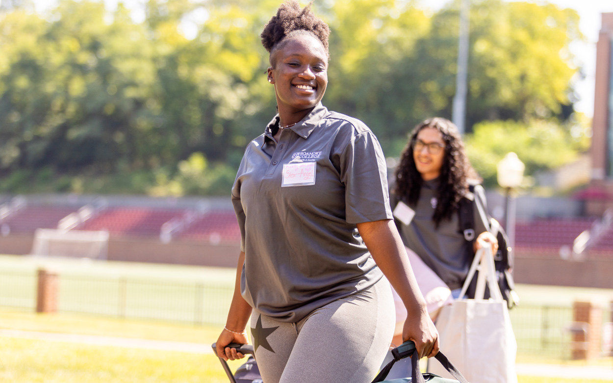 A student wearing a Roanoke College shirt smiles while helping carry bags into a residence hall