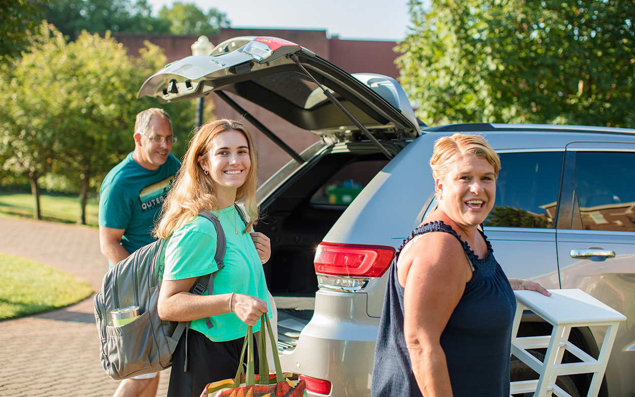 A student and her parents smile while unloading things from the trunk of their car