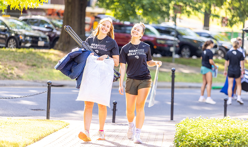 Two students wearing Roanoke Volleyball t-shirts smile while helping arriving students move their things into a residence hall