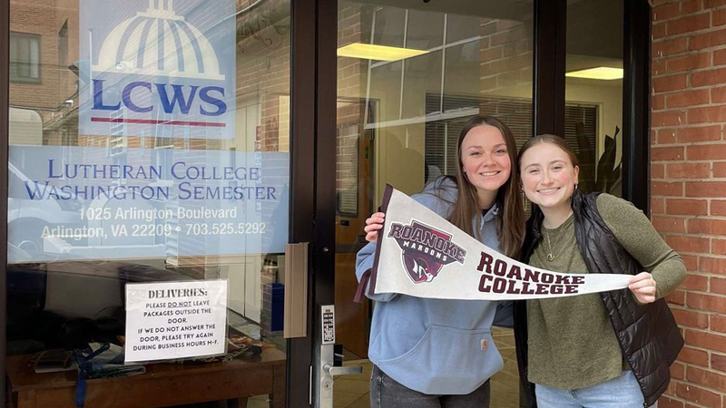 Heather Farnsworth and a fellow student smiling for a photo outside the Washington Semester offices while holding up a Roanoke College pennant