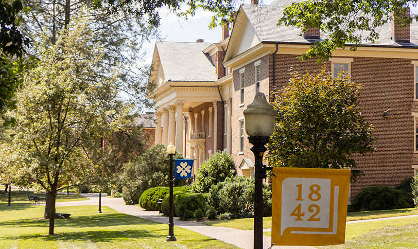 Brick buildings with white columns on a grassy campus, a yellow flag in the foreground.