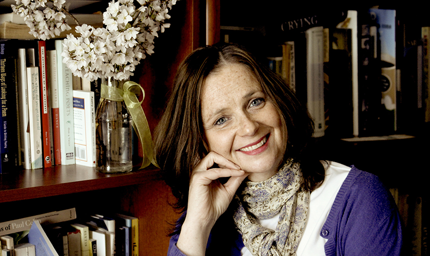 Headshot of a dark-haired woman in a purple sweater and cream scarf in front of a set of bookshelves that has books and a vase of white flowers on it. 