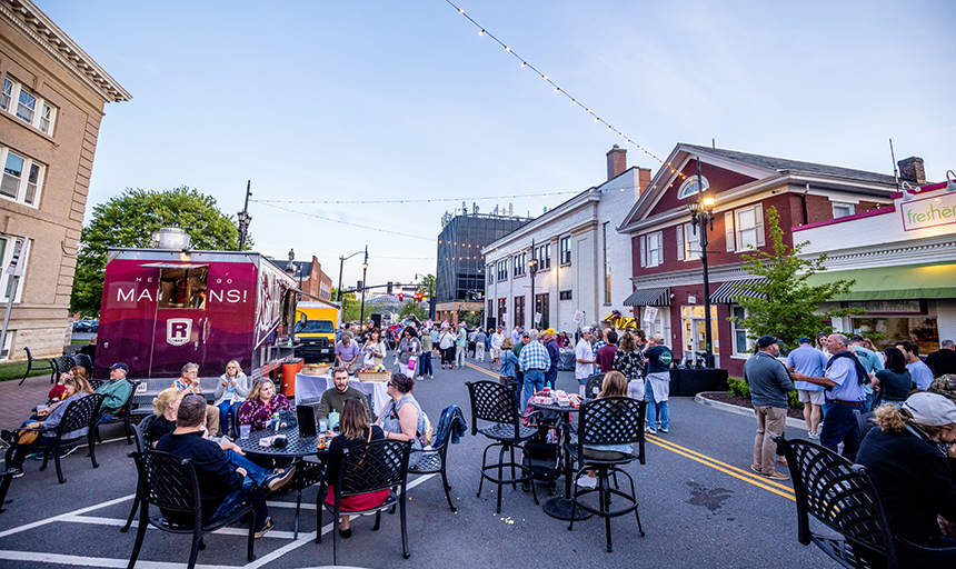 Groups of people sitting at tables and eating and talking. Tables are in the middle of a street with white lights strung across overhead.