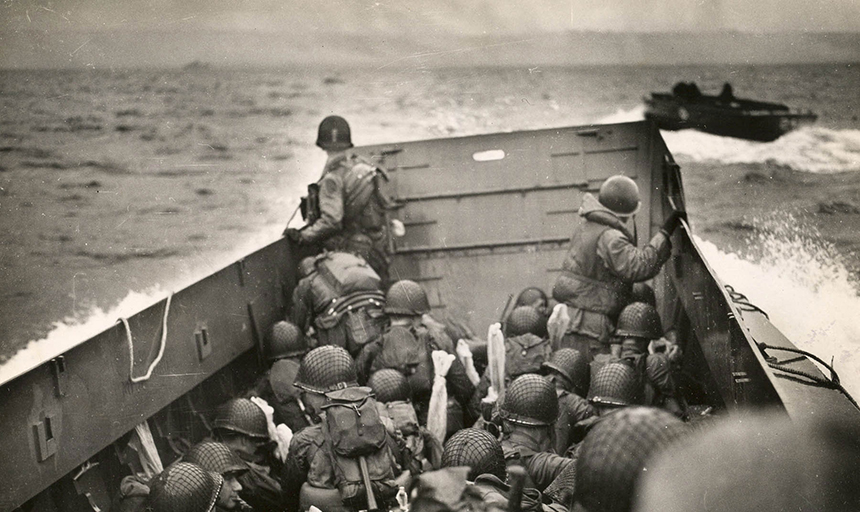 Black-and-white image showing helmeted servicemen crouching in a landing craft vehicle that is plowing across the ocean toward the coast of France. Public domain photo courtesy of 