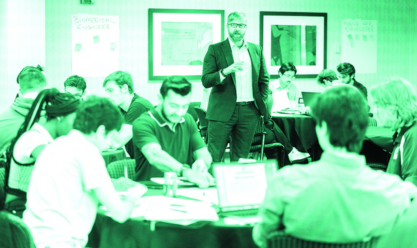 Green-tinted photo of a man standing among round tables where students are seated.