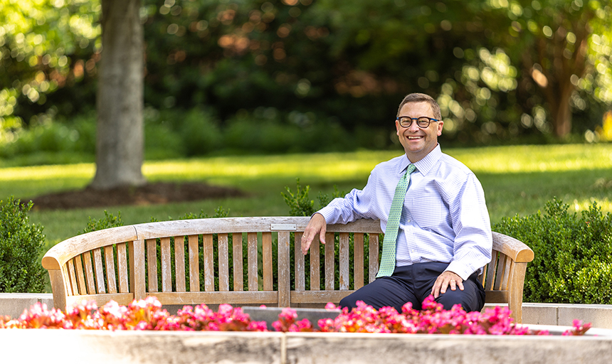 Man in a blue shirt and tie sits on a park bench with pink flowers in the foreground and green grass and trees behind him.